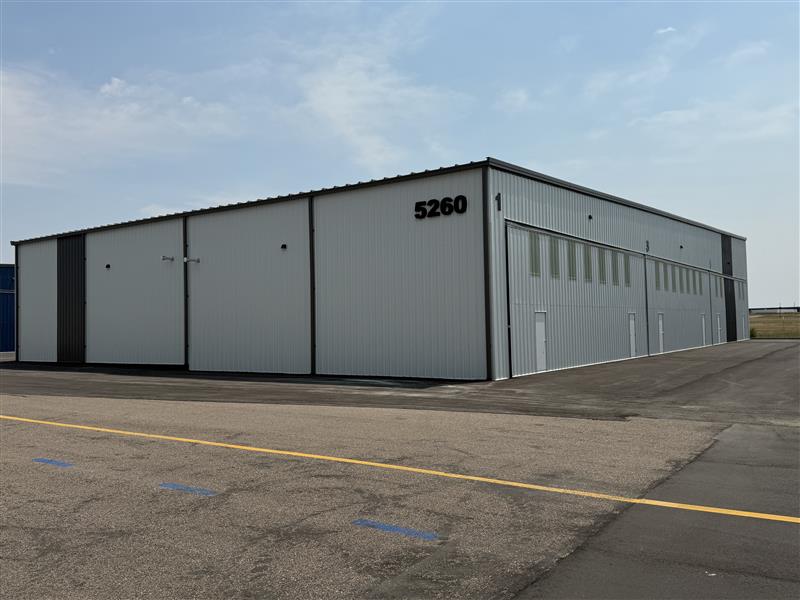 Business Aviation Group Large white industrial warehouse building numbered 5260, with multiple garage doors and windows, located on a paved lot at Northern Colorado Regional (FNL airport) under a partly cloudy sky.