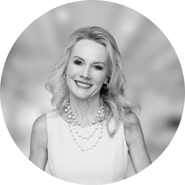 Business Aviation Group Black and white portrait of a smiling woman with wavy hair, wearing a sleeveless top and layered pearl necklaces, standing against a blurred background that hints at an aviation real estate brokerage office.