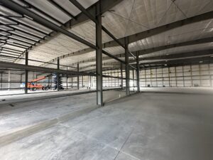 Business Aviation Group Interior of a large, unfinished warehouse by Tallyho Construction, featuring steel beams, concrete floor, and a scissor lift in the background; walls and ceiling are partially insulated.