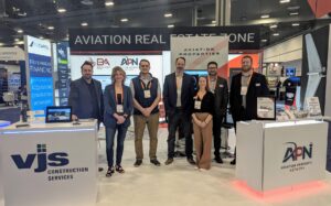 Business Aviation Group Group of seven people standing and posing for a photo at a trade show booth for VJS Construction Services and Aviation Property Network. Expo displays and signage visible in the background.