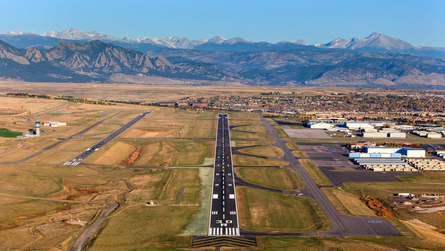 Business Aviation Group A wide view of Rocky Mountain Metropolitan Airport (BJC) shows the runway, taxiways, and buildings surrounded by open fields, with mountains in the background under a clear sky.